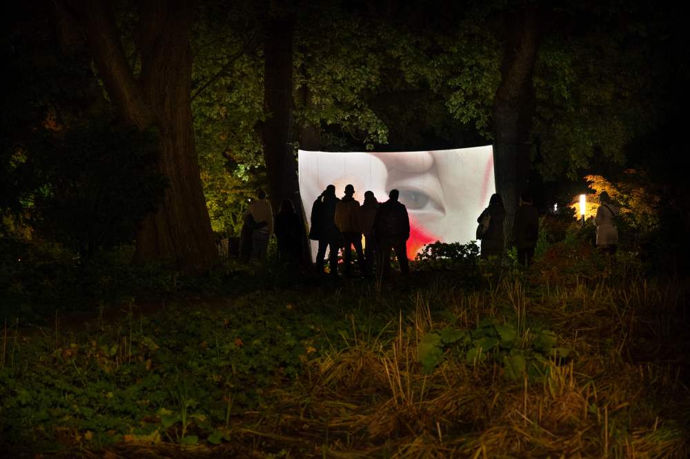 Installation at Planten un Blomen park in Hamburg by Hiền Hoàng, featuring large-scale video projections on towering trees. The immersive experience comprises three distinct screens, each showcasing a different film from the 'Scent from Heaven' project. The videos explore themes of tree’s suffering, human’s dream of healing and salvation and sensory perception, enveloping viewers in a mesmerizing blend of sight and sound amidst the natural surroundings of the park.
